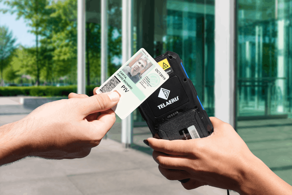 Hand holding a PIV card with photo ID up to a Telaeris handheld badge reader for scanning